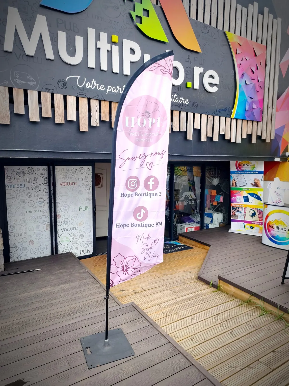 Drapeau personnalisé rose « Hope prêt-à-porter » sur son socle, devant la boutique MultiPub au Tampon